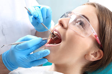 Dentist wearing blue gloves and safety glasses probes a patient's open mouth with dental instruments.