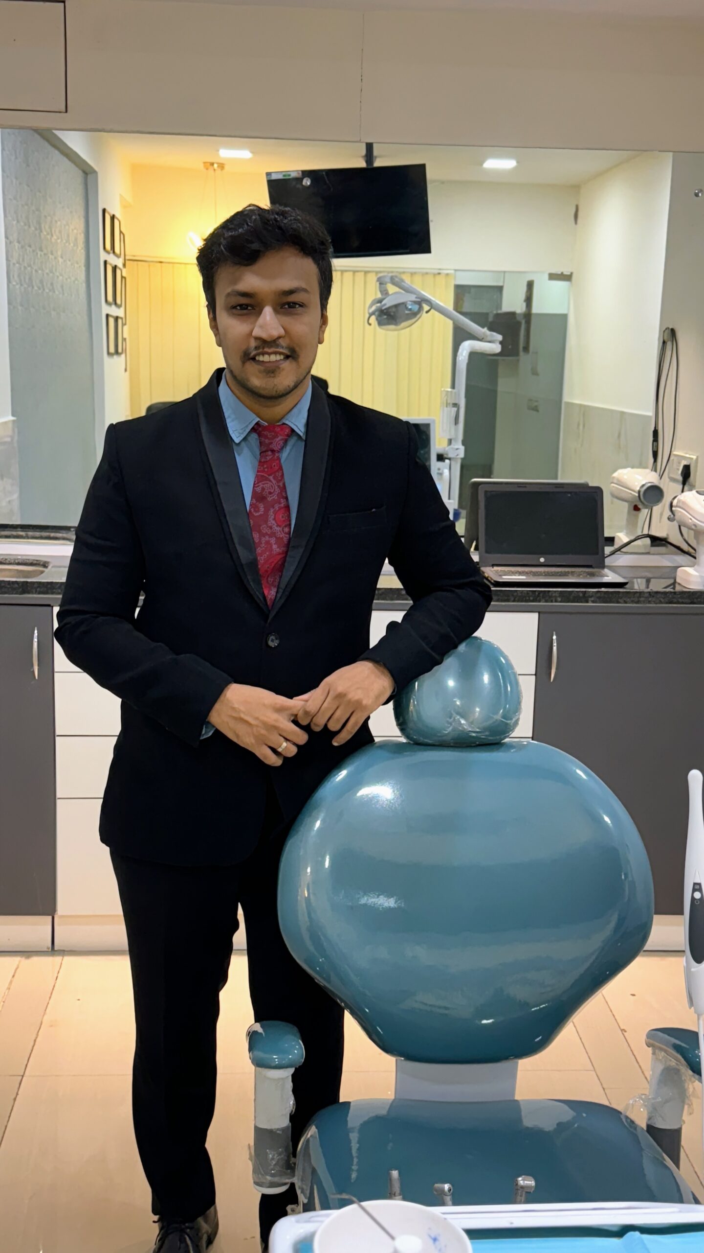 Man in a dark suit and red tie standing beside a large blue dental chair in a clinic setting.