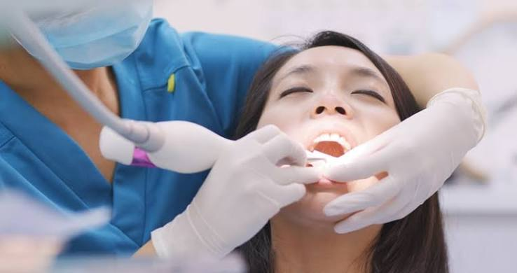 Dentist in blue scrubs and gloves uses a dental drill on a patient with an open mouth.