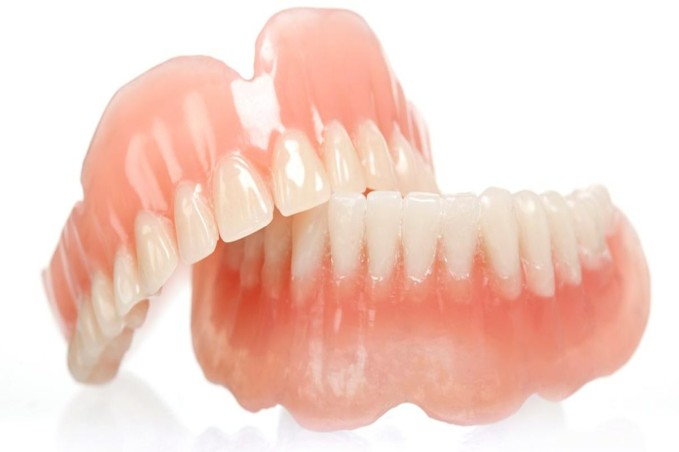 Complete-Dentures 1 - The Smile Connect Full set of removable dentures with pink acrylic gums and white teeth on a white background/reflective surface